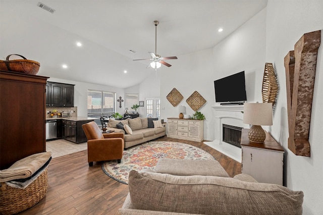 living area with visible vents, a fireplace with raised hearth, ceiling fan, high vaulted ceiling, and light wood-type flooring