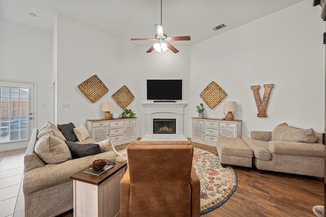 living room featuring visible vents, a fireplace with raised hearth, a high ceiling, dark wood-type flooring, and ceiling fan