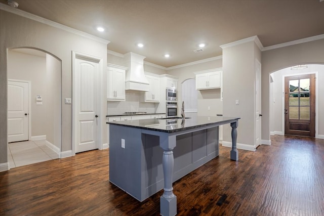 kitchen with arched walkways, appliances with stainless steel finishes, an island with sink, and custom exhaust hood