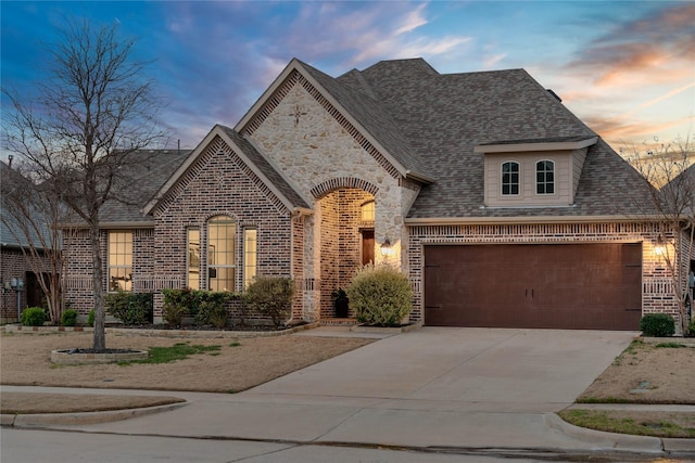 french country style house with a garage, a shingled roof, concrete driveway, stone siding, and brick siding