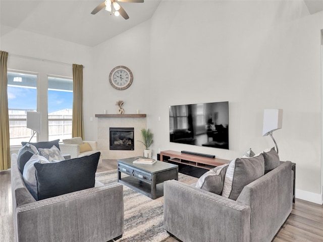 living area featuring ceiling fan, wood finished floors, high vaulted ceiling, a tile fireplace, and baseboards