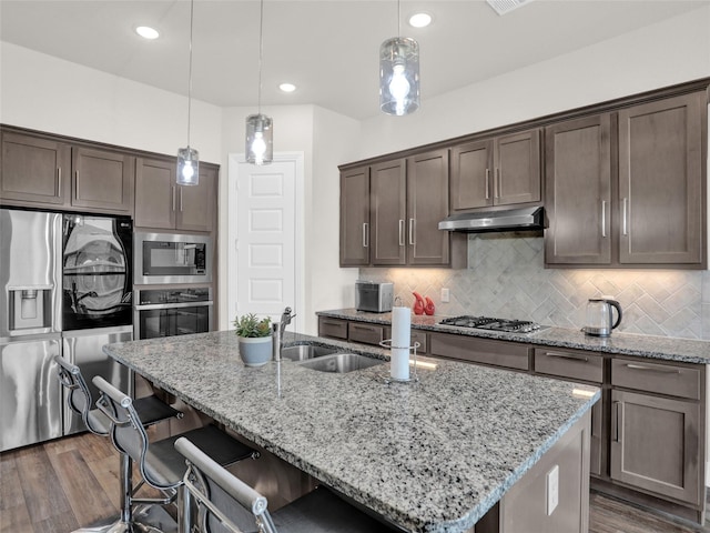 kitchen with dark brown cabinetry, under cabinet range hood, stainless steel appliances, an island with sink, and dark wood finished floors