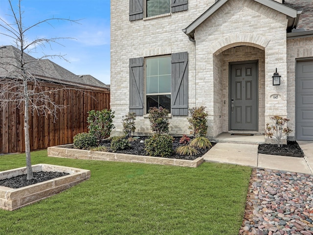 view of exterior entry with brick siding, a lawn, an attached garage, and fence