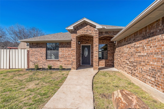 doorway to property with a shingled roof, brick siding, a lawn, and fence