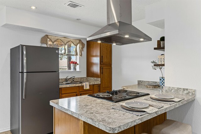 kitchen featuring black electric stovetop, a peninsula, island exhaust hood, freestanding refrigerator, and brown cabinetry