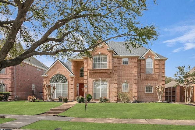 traditional-style house featuring a front lawn, roof with shingles, fence, and brick siding