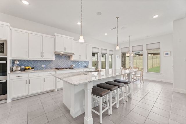 kitchen with light tile patterned floors, under cabinet range hood, a sink, backsplash, and stainless steel gas stovetop
