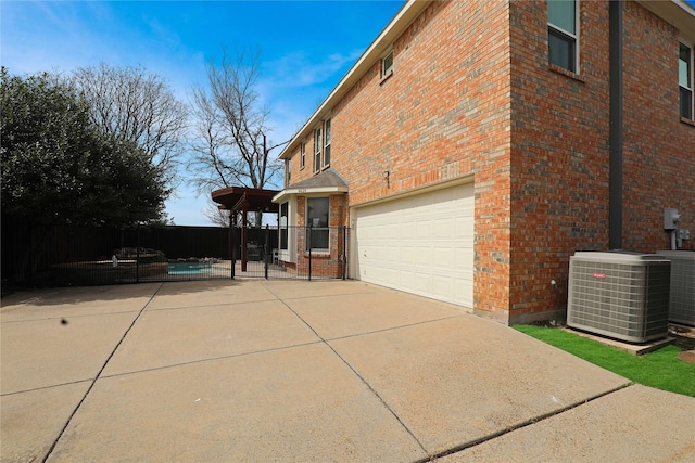 view of side of property featuring driveway, fence, an attached garage, brick siding, and central AC unit