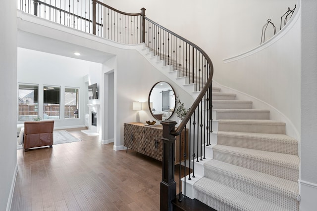 staircase with a glass covered fireplace, a towering ceiling, baseboards, and wood finished floors