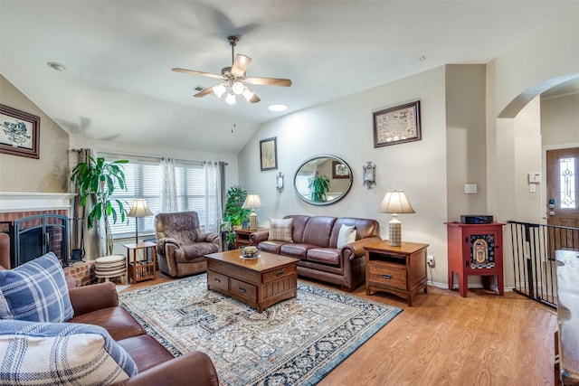 living room with lofted ceiling, arched walkways, light wood-style flooring, a ceiling fan, and a brick fireplace