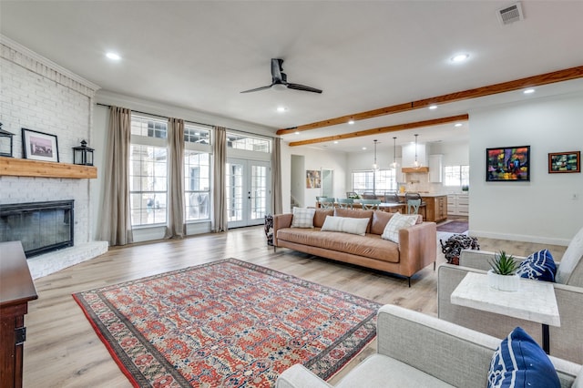 living room featuring a ceiling fan, beamed ceiling, visible vents, light wood-style flooring, and a brick fireplace