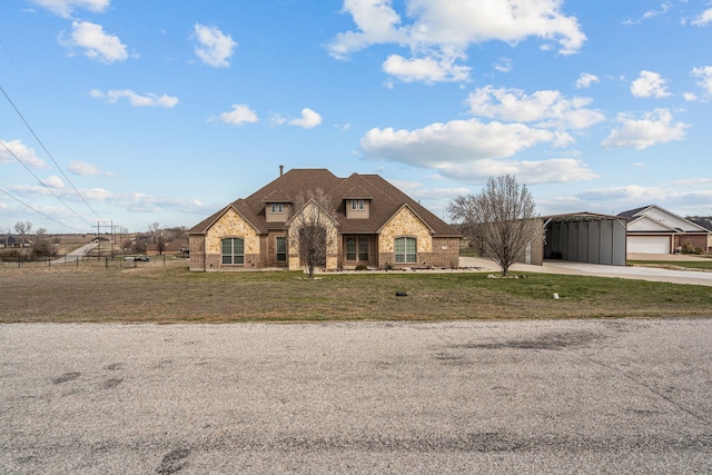 french country home with a shingled roof, a detached carport, a front yard, stone siding, and an outdoor structure