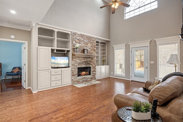 living area with baseboards, a ceiling fan, wood finished floors, crown molding, and a stone fireplace