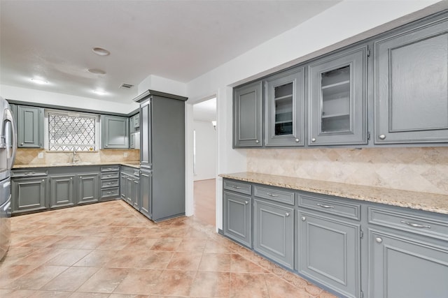 kitchen with gray cabinetry, a sink, light stone countertops, tasteful backsplash, and glass insert cabinets