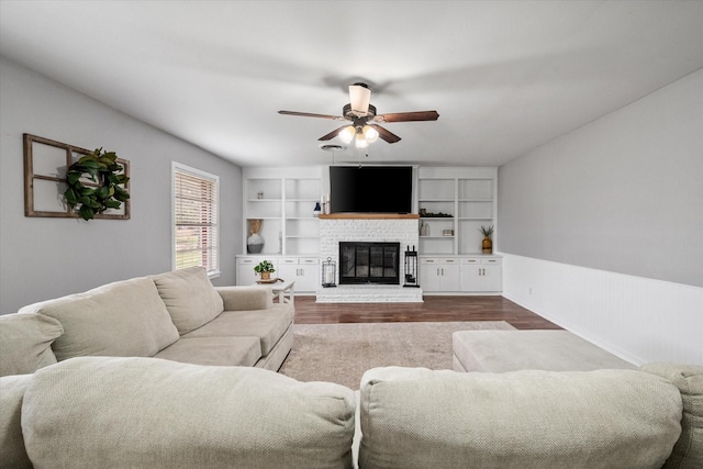 living room with a brick fireplace, ceiling fan, built in shelves, and dark wood-type flooring