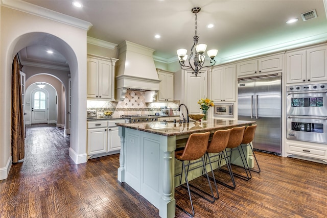 kitchen with arched walkways, dark wood finished floors, built in appliances, custom exhaust hood, and backsplash