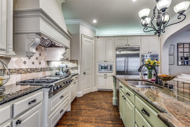 kitchen with arched walkways, dark stone countertops, built in appliances, a sink, and green cabinetry