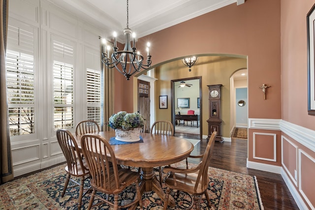 dining space featuring arched walkways, dark wood-style flooring, crown molding, a decorative wall, and ceiling fan with notable chandelier