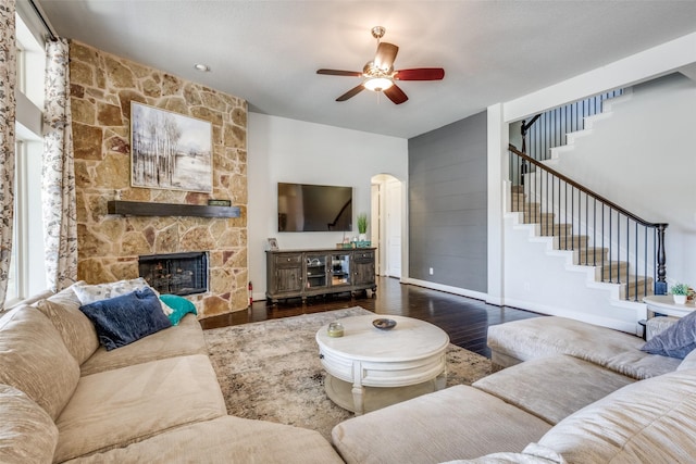 living room with arched walkways, a stone fireplace, wood finished floors, baseboards, and stairs