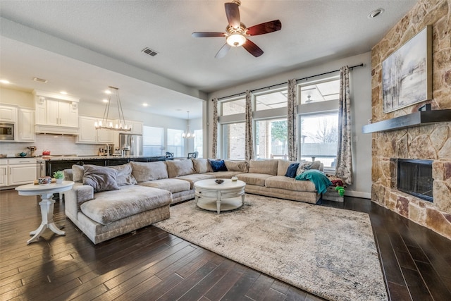 living area with dark wood-style flooring, visible vents, a stone fireplace, a textured ceiling, and ceiling fan with notable chandelier