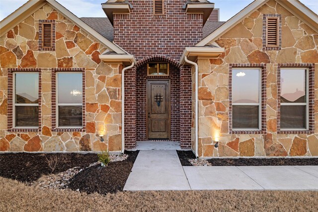 doorway to property featuring stone siding, brick siding, and roof with shingles