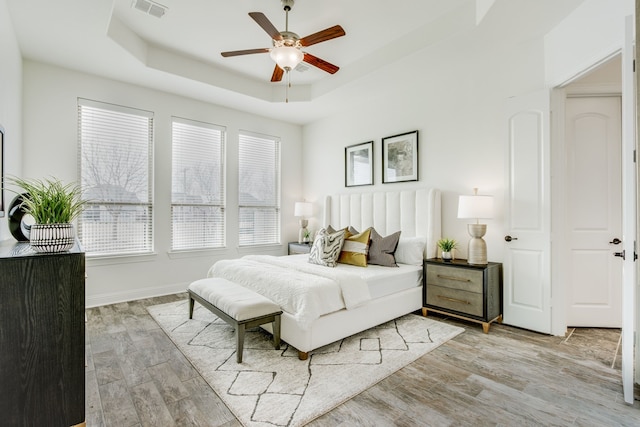 bedroom featuring a tray ceiling, light wood finished floors, visible vents, a ceiling fan, and baseboards