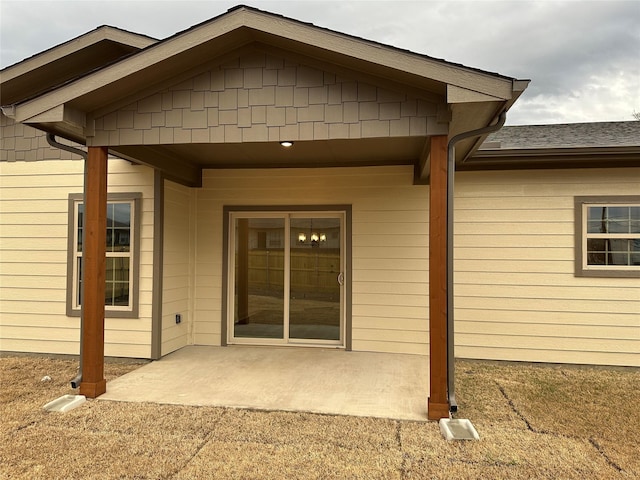 view of exterior entry featuring roof with shingles and a patio