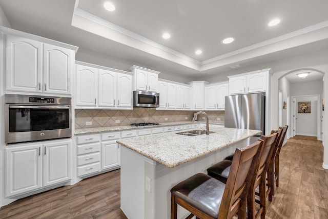 kitchen featuring a tray ceiling, arched walkways, stainless steel appliances, backsplash, and a sink