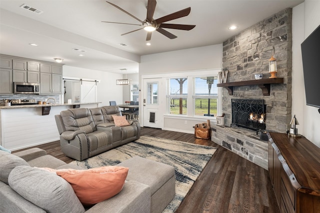 living area with a barn door, visible vents, ceiling fan, dark wood-type flooring, and a stone fireplace