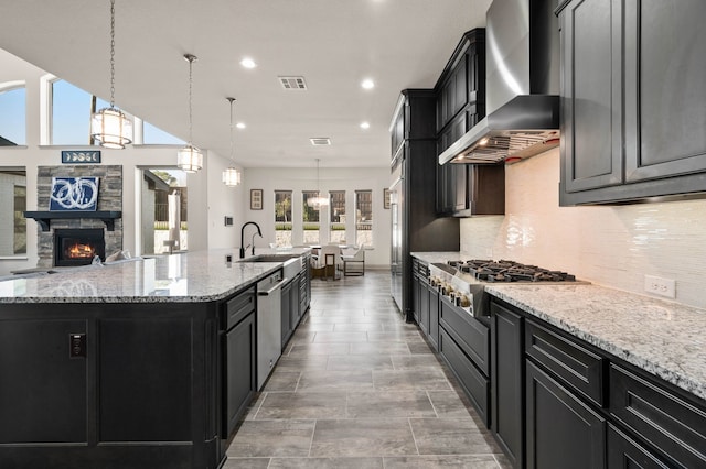 kitchen with dark cabinets, stainless steel appliances, visible vents, wall chimney exhaust hood, and a large island with sink