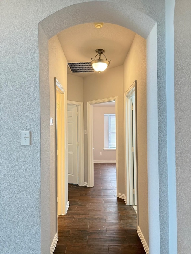 corridor featuring arched walkways, visible vents, a textured wall, dark wood-type flooring, and baseboards