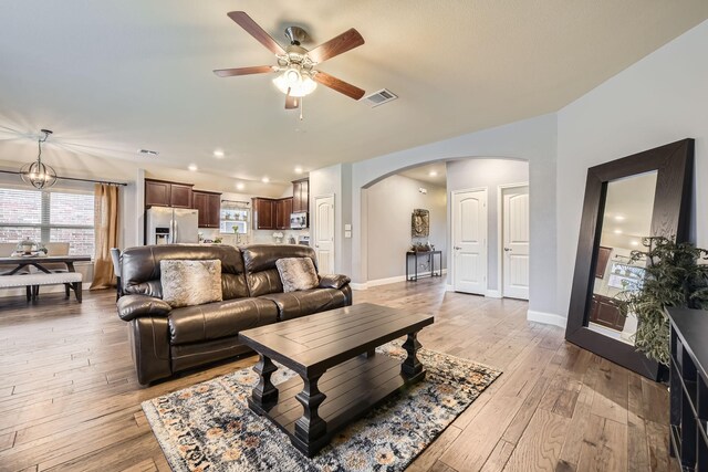 living area with visible vents, baseboards, light wood-style flooring, ceiling fan with notable chandelier, and arched walkways