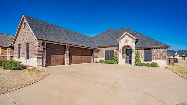 french country inspired facade with roof with shingles, brick siding, an attached garage, stone siding, and driveway