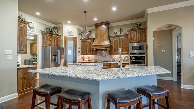 kitchen with arched walkways, stainless steel appliances, custom range hood, a sink, and a large island with sink