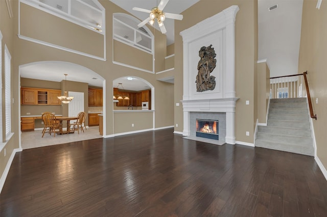 unfurnished living room featuring ceiling fan with notable chandelier, dark wood-type flooring, visible vents, stairs, and a tiled fireplace