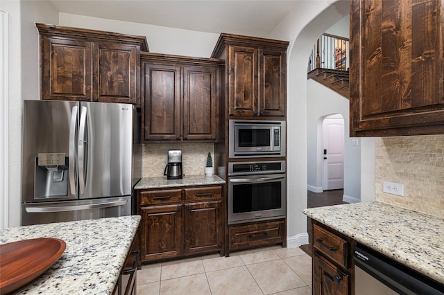 kitchen featuring arched walkways, light stone counters, dark brown cabinets, appliances with stainless steel finishes, and tasteful backsplash