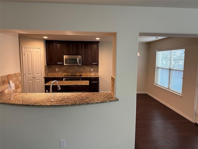 kitchen featuring baseboards, decorative backsplash, stainless steel microwave, light stone countertops, and dark brown cabinets