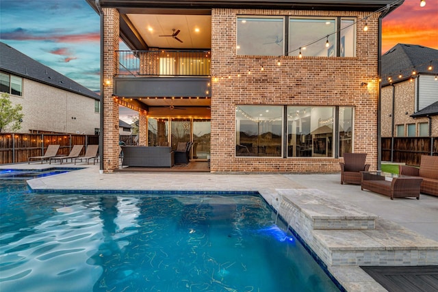 back of house at dusk with a fenced in pool, a balcony, fence, and brick siding