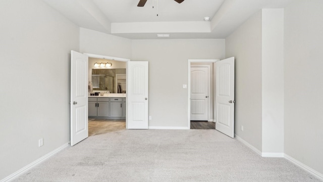 unfurnished bedroom with light carpet, a raised ceiling, connected bathroom, baseboards, and ceiling fan