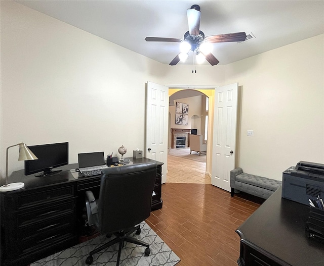 office with arched walkways, a glass covered fireplace, a ceiling fan, and wood finished floors