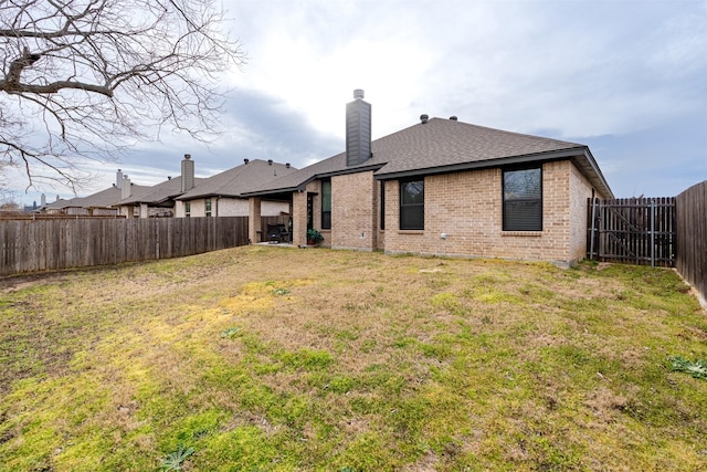 back of house with a fenced backyard, brick siding, a shingled roof, a lawn, and a chimney