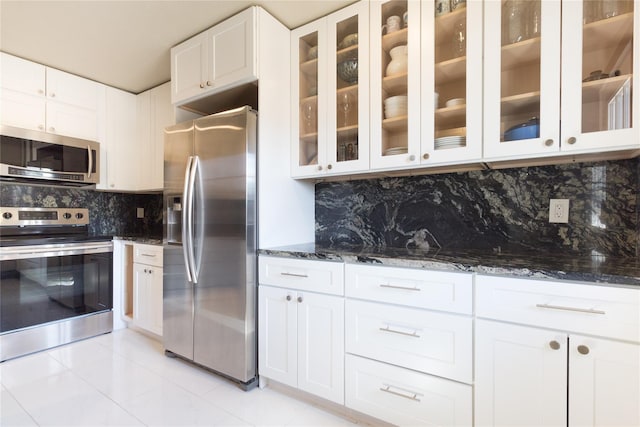 kitchen with white cabinets, appliances with stainless steel finishes, backsplash, dark stone countertops, and glass insert cabinets