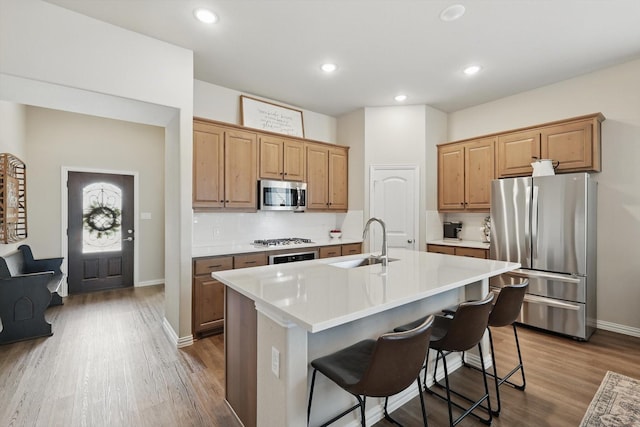 kitchen with a kitchen island with sink, stainless steel appliances, wood finished floors, a sink, and light countertops