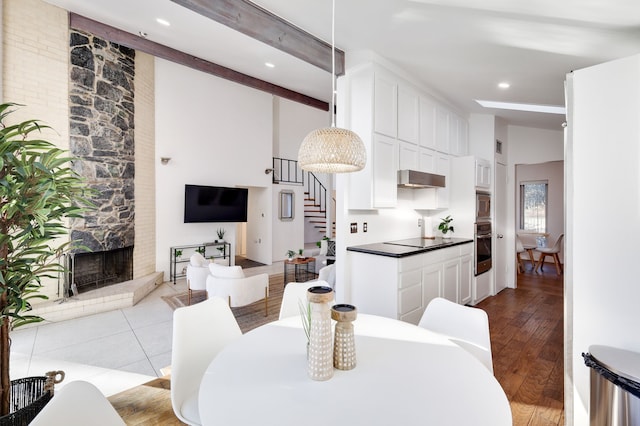 dining room with a towering ceiling, wood finished floors, stairs, a stone fireplace, and beam ceiling