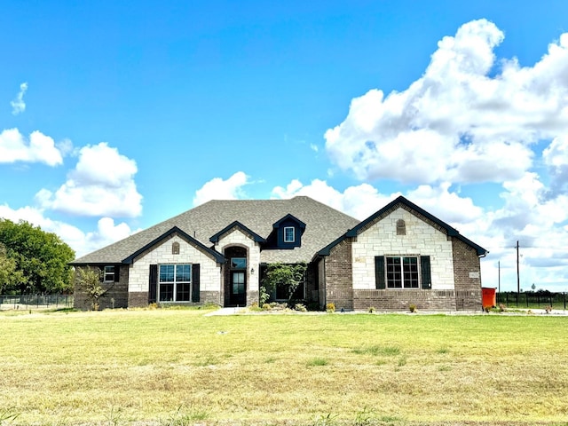 french country style house with stone siding, a shingled roof, a front yard, and fence