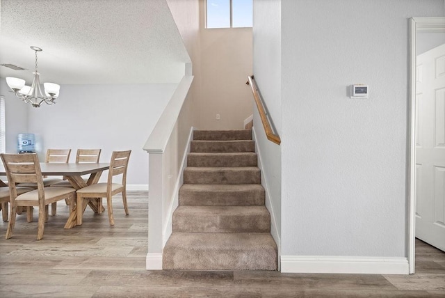 staircase with a textured ceiling, a notable chandelier, wood finished floors, visible vents, and baseboards