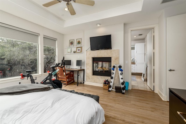 bedroom featuring a raised ceiling, light wood-style flooring, a tiled fireplace, ceiling fan, and baseboards