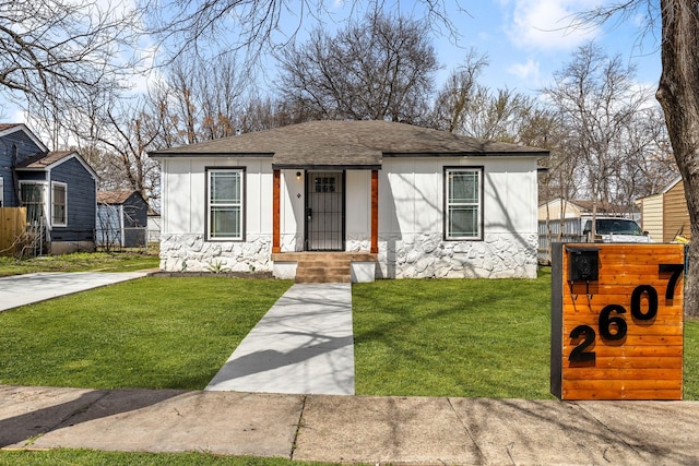 bungalow featuring stone siding, a front lawn, roof with shingles, and fence