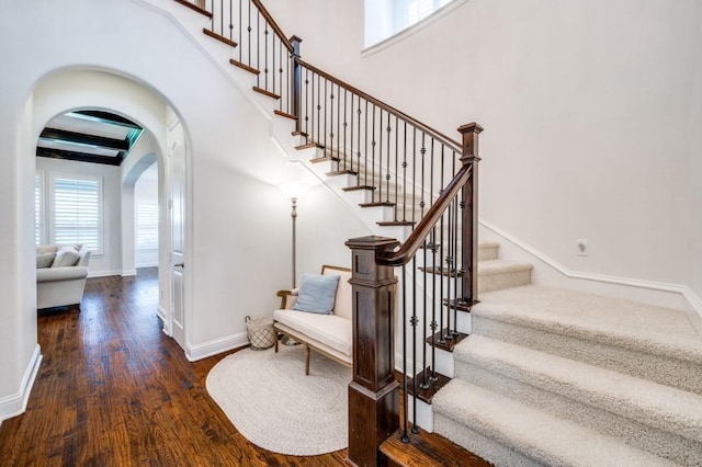 stairs featuring arched walkways, wood finished floors, a towering ceiling, baseboards, and beam ceiling