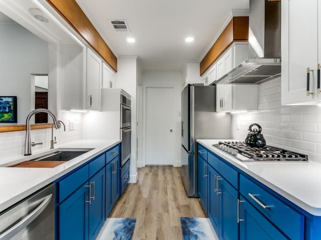 kitchen with blue cabinets, a sink, white cabinetry, stainless steel appliances, and wall chimney range hood